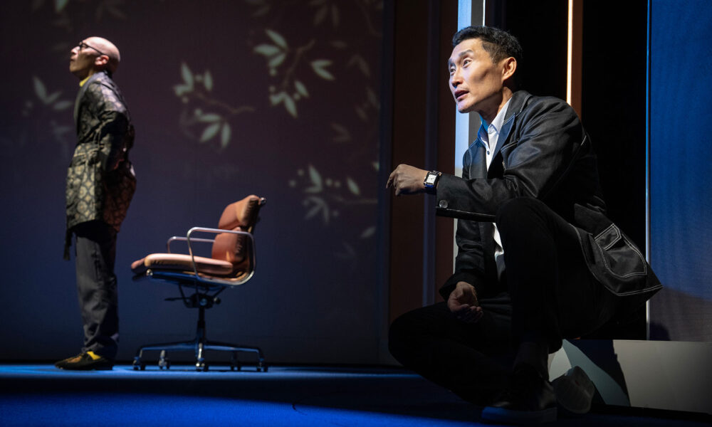 Asian man in a suit kneeling and looking to the side, in the background is a bald Asian man standing